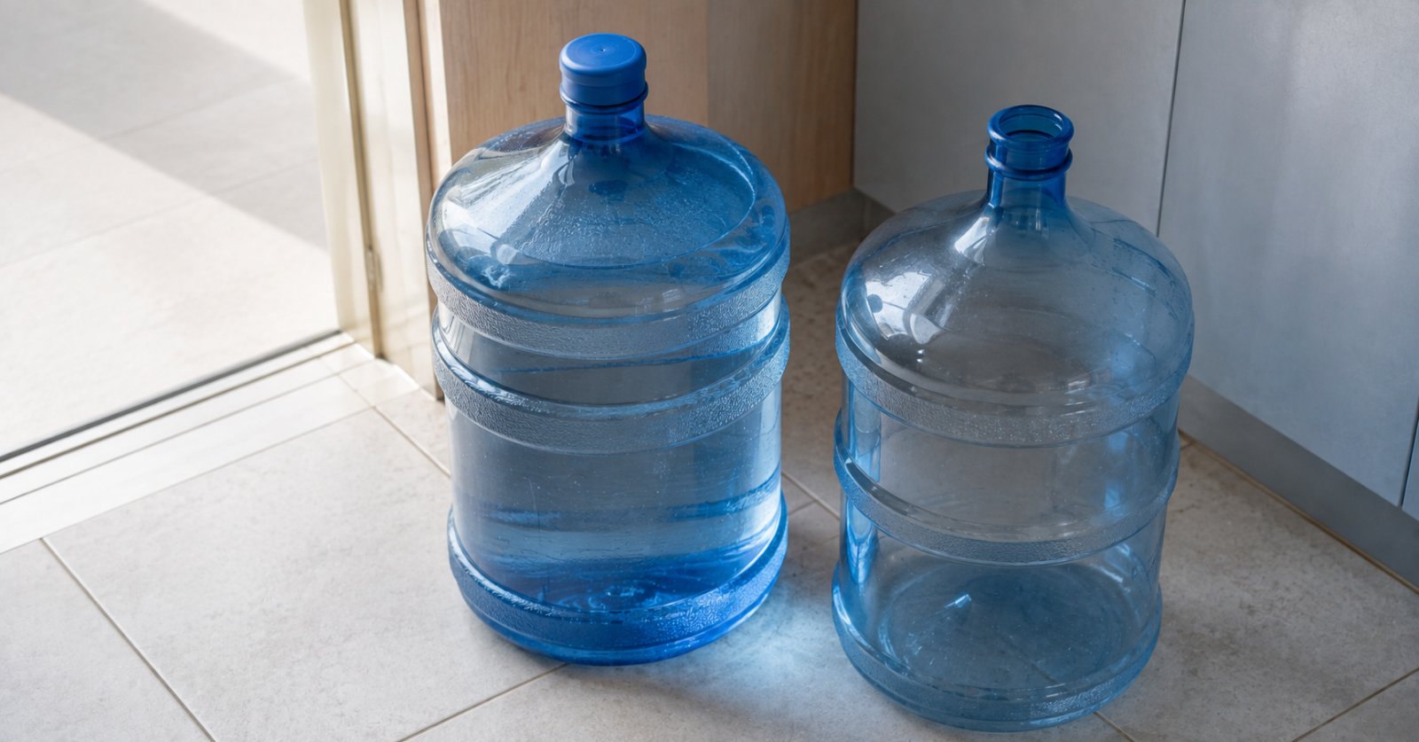 Dos botellones de agua sobre el piso de una cocina, uno lleno y uno vacío